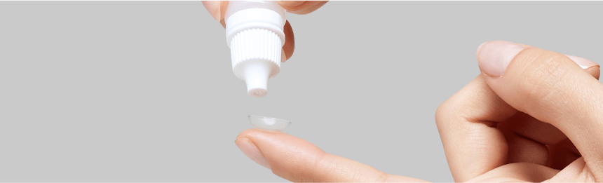 Hand holding a soft lens on a fingertip while a bottle dispenses cleaning solution, illustrating proper contact lenses hygiene.
